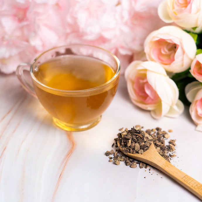 Cup of Organic Black Cohosh tea with dried roots on a wooden spoon and pink flowers in the background.