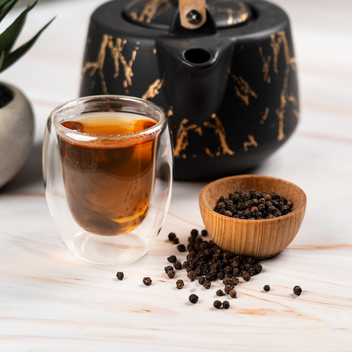 Glass teacup filled with black pepper tea beside a small wooden bowl of whole black peppercorns on a light wooden surface, with a black and gold teapot in the background.