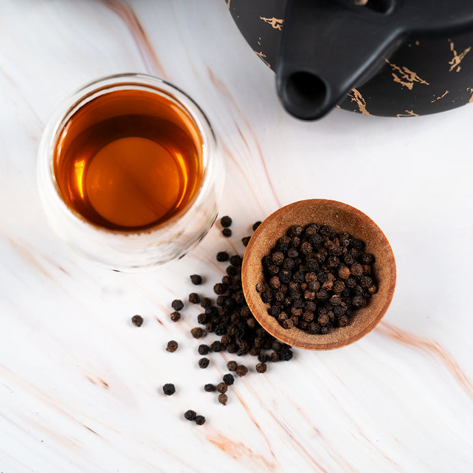 Cup of brewed tea beside a bowl of black peppercorns and a teapot on a marble surface.