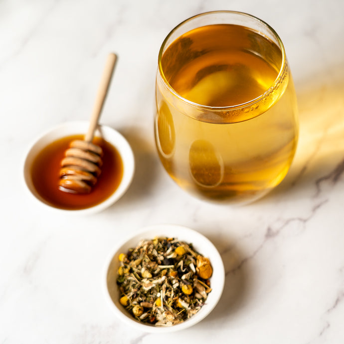 Glass of Organic Bladder Health Tea with a bowl of loose herbal blend and honey on a marble surface.