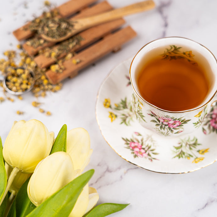 Cup of Organic Bloat Relief Tea in a floral teacup beside loose tea, a wooden scoop, and yellow tulips on a marble surface.