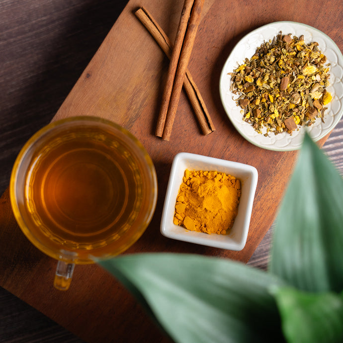 A top view of Organic Blood Sugar Balance Tea in a glass cup on a wooden board, styled with cinnamon sticks, a small dish of loose leaf tea, and a square bowl of golden turmeric powder.