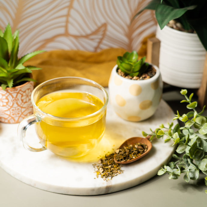 A clear glass mug of bright yellow Organic Brain Health Tea on a marble tray, surrounded by small potted plants and a wooden spoon with loose leaf tea.