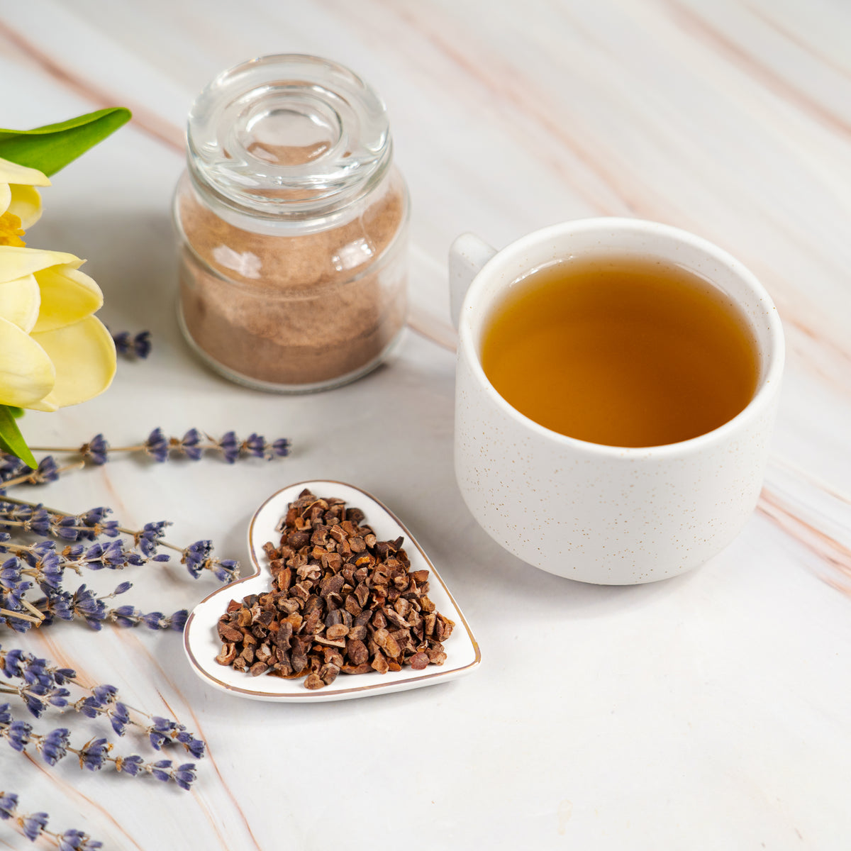 White ceramic mug filled with cacao nib tea beside a small glass jar of cocoa powder and a heart-shaped dish of cacao nibs, decorated with sprigs of lavender and yellow tulip petals on a white marble surface.