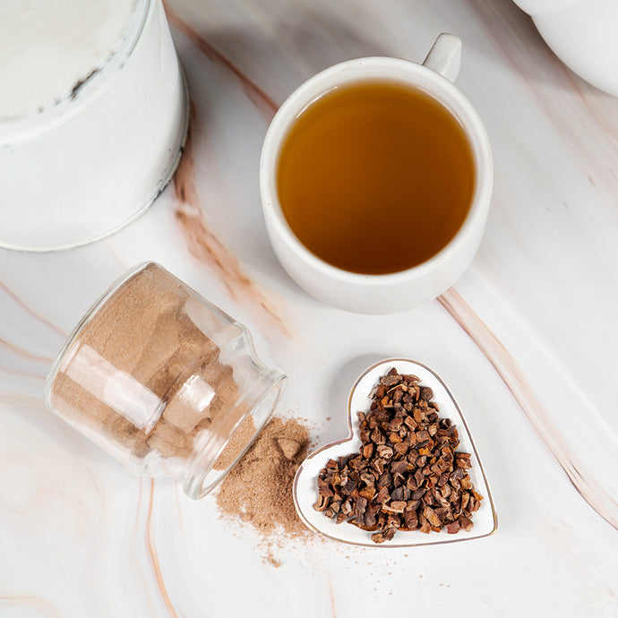 Tea cup with a heart-shaped container of cacao nibs and a jar of ground cacao powder on a marble surface.