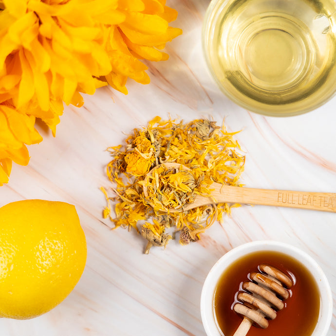 Tea-making ingredients including dried calendula flowers, a wooden spoon, a cup of tea, a honeycomb, and a lemon on a white surface.