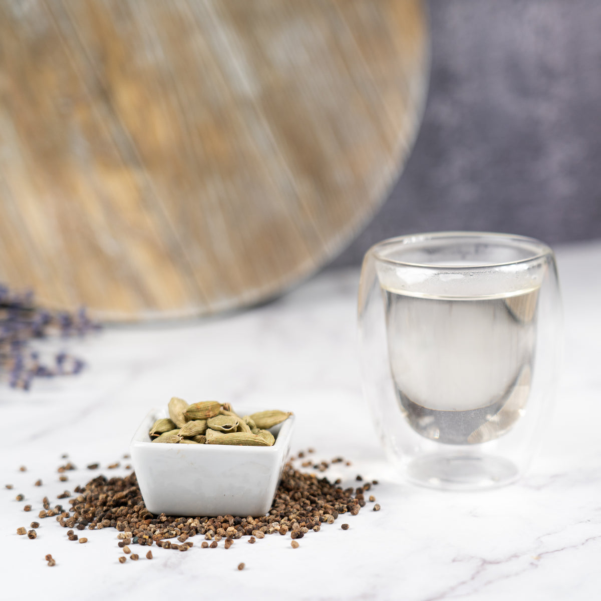 Small white dish filled with green cardamom pods on a marble surface beside scattered cardamom seeds and a clear glass cup of hot water, with a wooden board in the background.