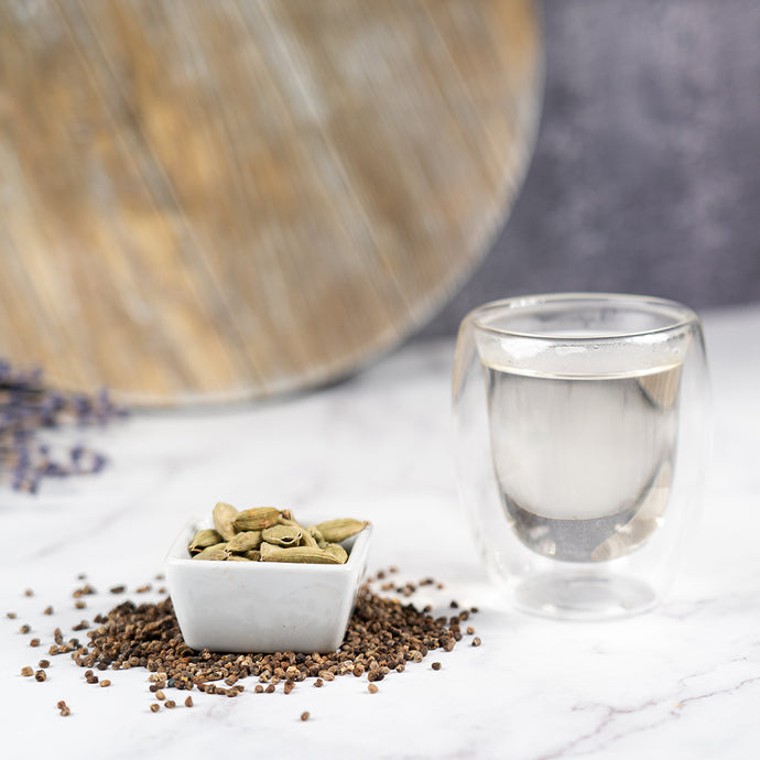 Cardamon pods in a small white dish with a glass of water on a marble surface.