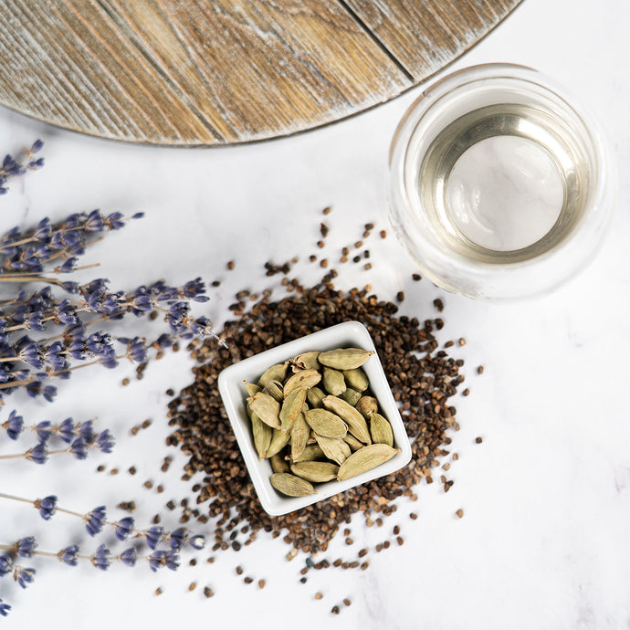 Cardamom pods in a white container with lavender flowers and a wooden board on a white surface.