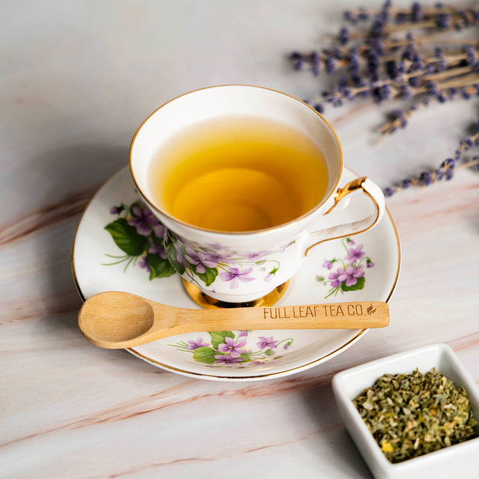 Tea cup with floral design on a saucer, wooden spoon, and catnip leaves on a marble surface.