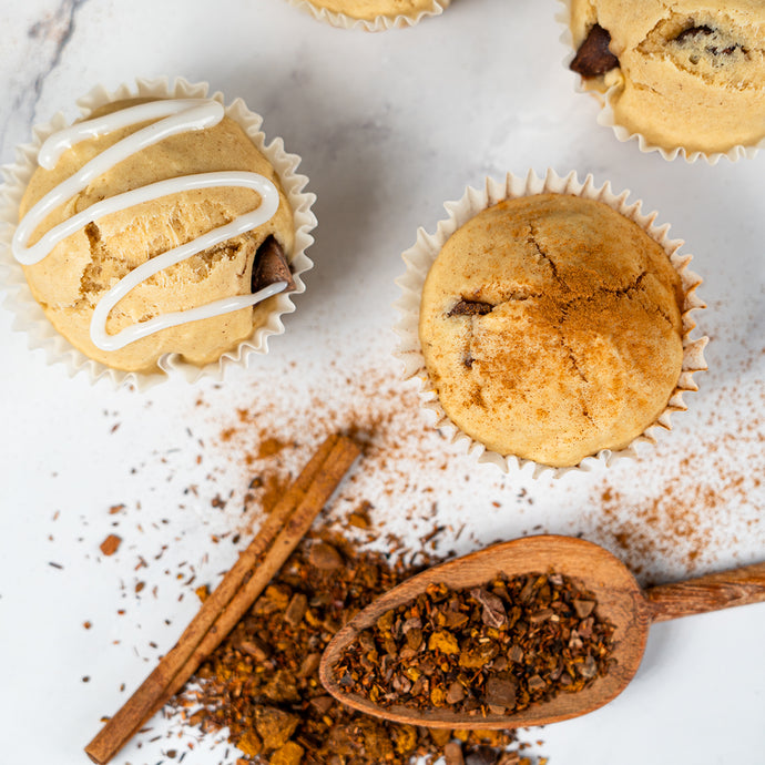 Muffins with white drizzle, cinnamon sticks, and a wooden spoon with chaga spices on a marble surface.