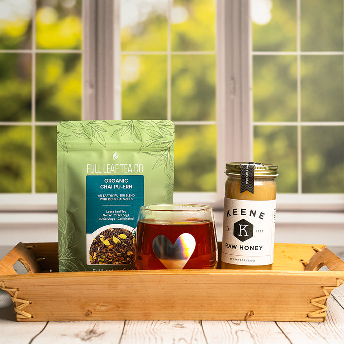Full leaf tea package of Organic chai pu-erh, glass of tea, and jar of honey on a wooden tray with a blurred green outdoor background.