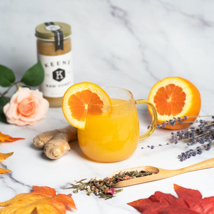 Glass mug with orange slices, jar of honey, ginger root, and lavender on a marble surface.