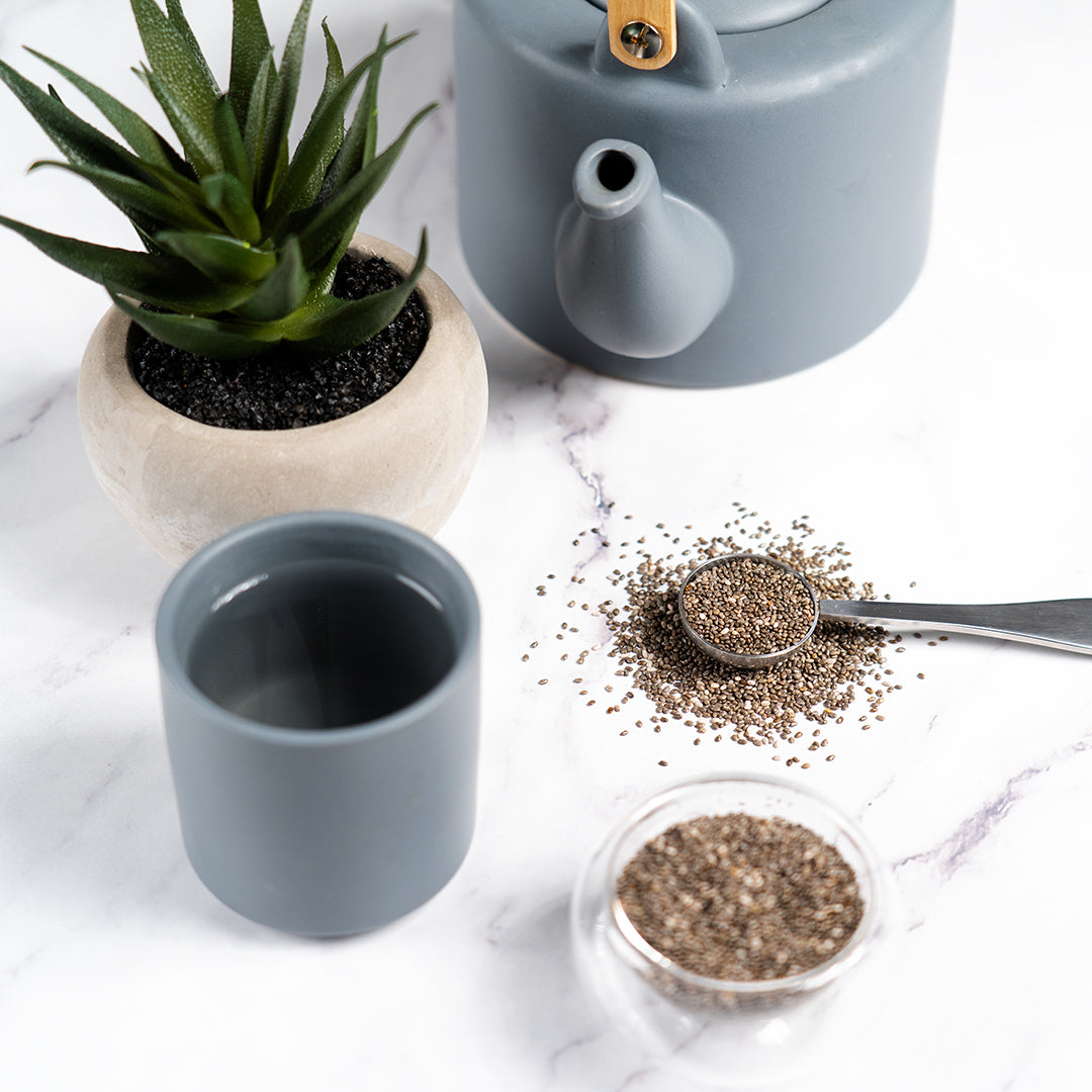 Tea set with gray teapot, cup, and small bowl of chia seeds on a marble surface.