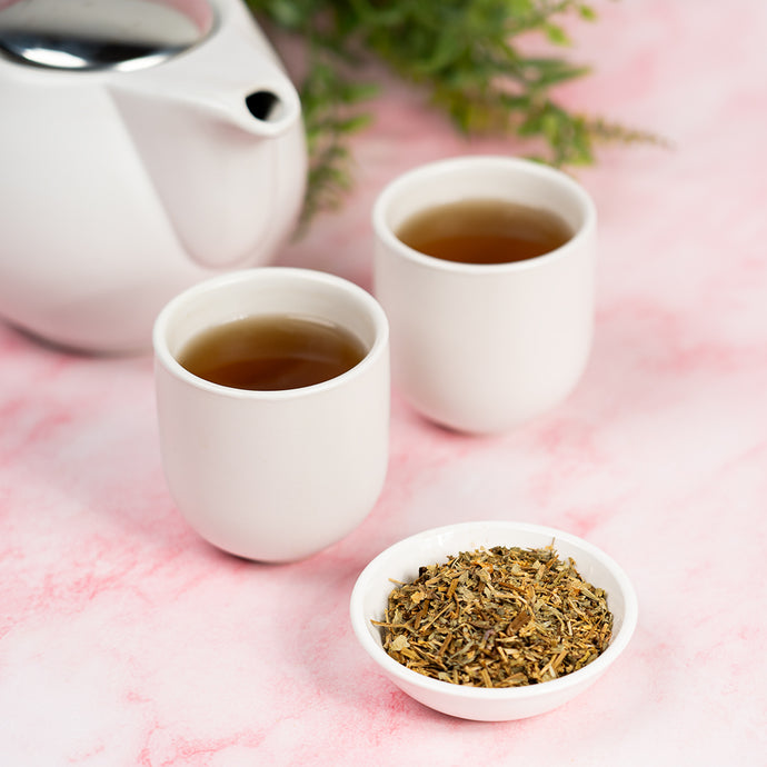 Two cups of tea with a teapot and a bowl of chickweed on a pink surface.
