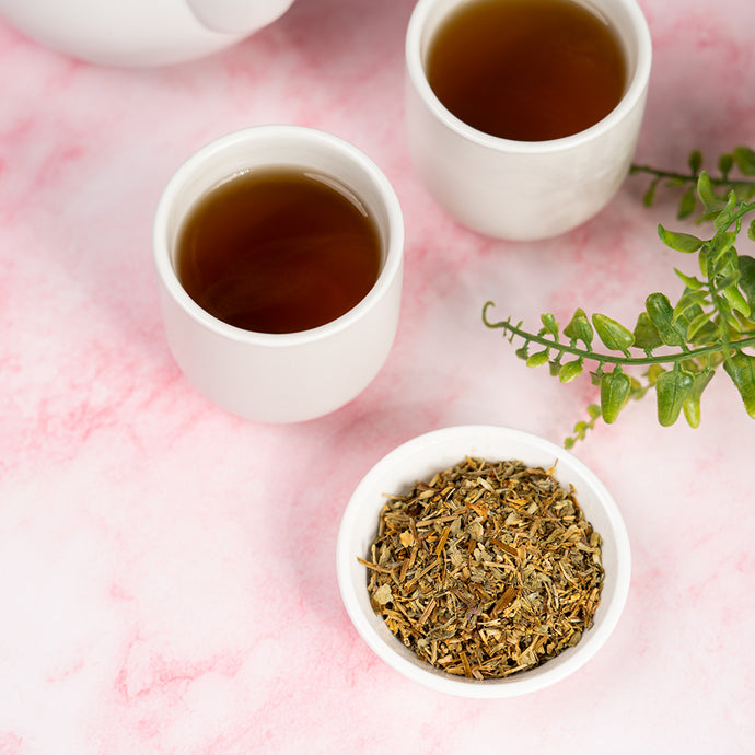 Two cups of tea and a bowl of dried chickweed on a pink surface with green leaves.