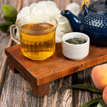 Glass cup of freshly brewed green tea with a blue teapot and a small bowl of loose leaf tea on a wooden tray, surrounded by white flowers and peaches.