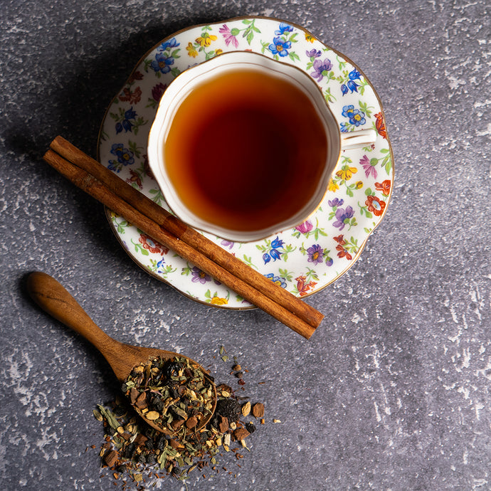 Teacup with floral saucer, cinnamon sticks, and a wooden spoon with tea leaves on a gray surface.