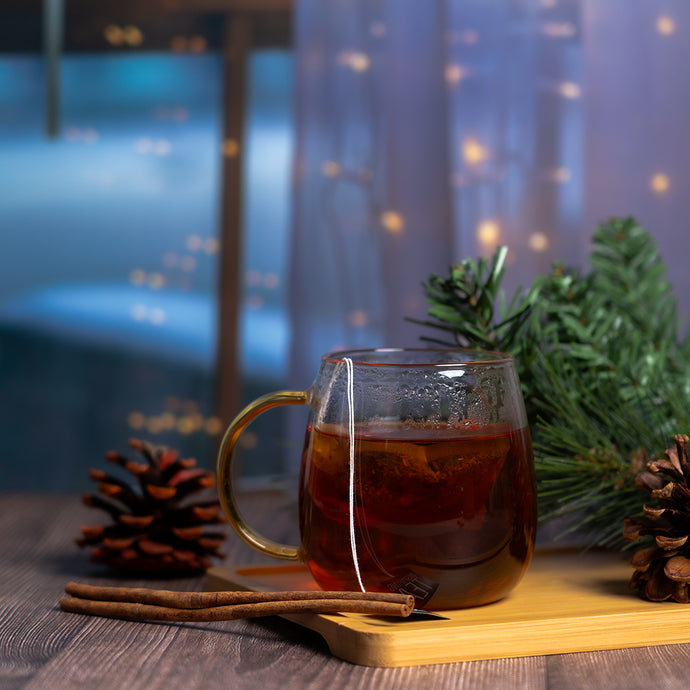 Glass mug with hot tea, cinnamon sticks, and pine cones on a wooden surface with a blurred festive background.