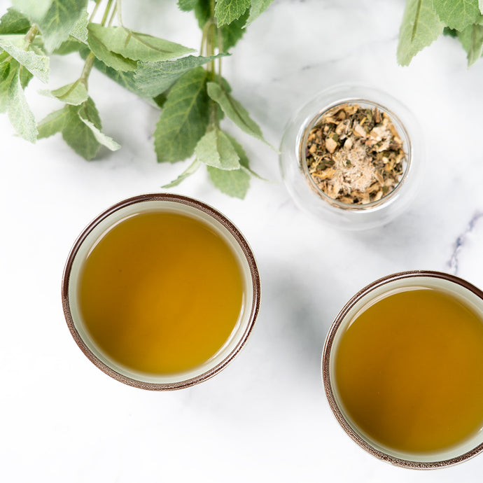Two cups of tea with a small container of herbs on a white surface.