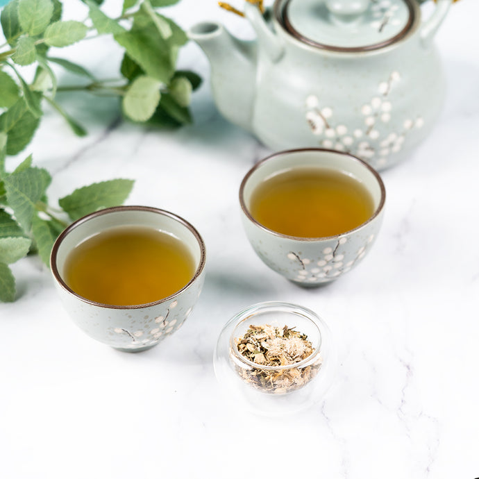 Two cups of cough relief tea on a marble surface with a tea pot and leaves in the background. 