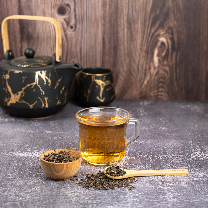 Tea set with black teapot, glass of tea, wooden spoon, and darjeeling tea leaves on a dark surface.