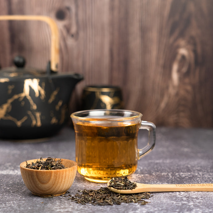 Glass cup of tea with a teapot, wooden spoon, and bowl of tea leaves on a wooden surface.