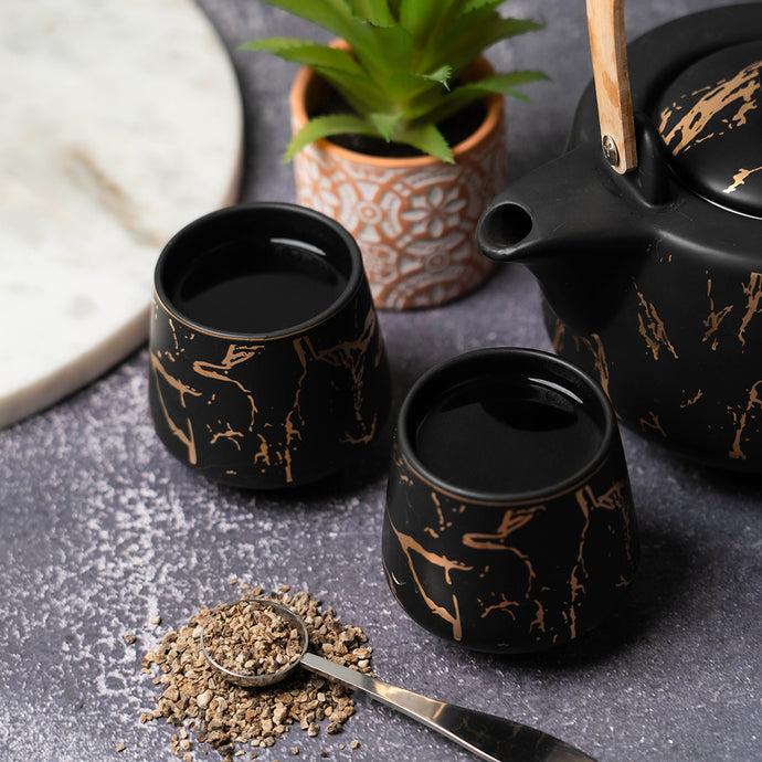 Black marbled teacups and teapot with devil's claw tea leaves and spoon on a dark surface