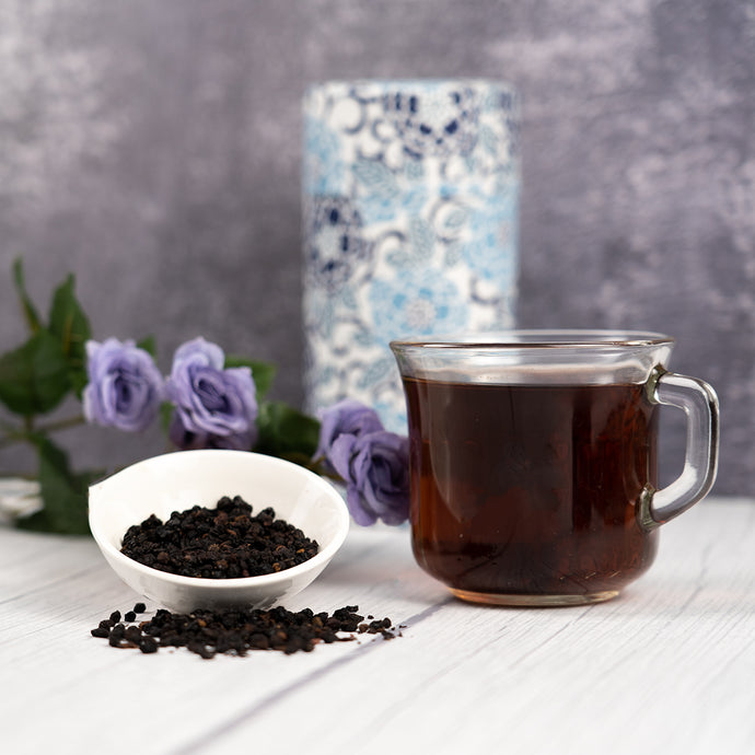 Clear glass mug filled with elderberry tea, surrounded by a bowl of black tea leaves and purple flowers on a wooden surface.
