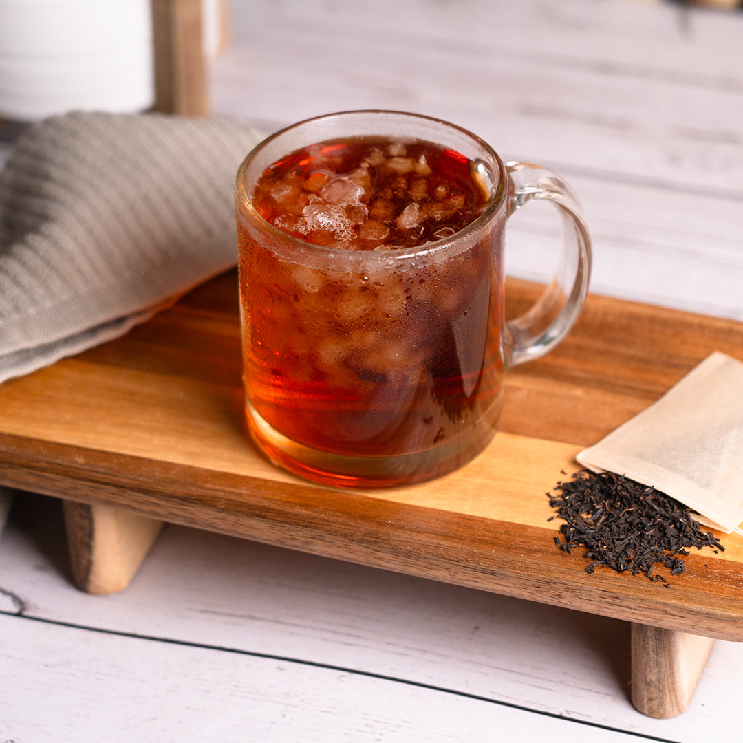 A glass mug filled with iced Organic English Breakfast sits on a wooden tray, surrounded by loose black tea leaves and a tea bag. The drink has a deep amber color with ice floating on top, and a folded gray cloth is placed nearby on a light wooden surface.