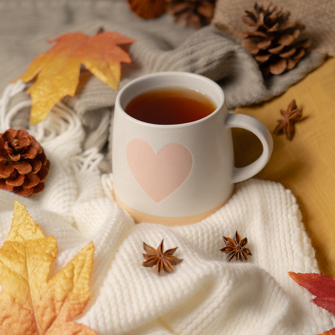 White mug with a pink heart on a white blanket surrounded by autumn leaves, pine cones, and star anise.