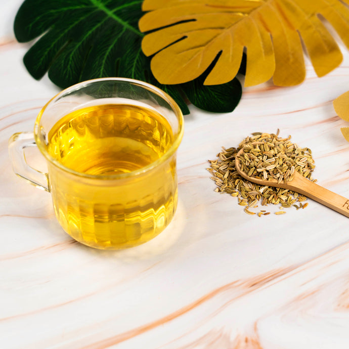Glass cup of Organic Fennel tea with wooden spoon and fennel seeds on a light marble surface with decorative leaves.