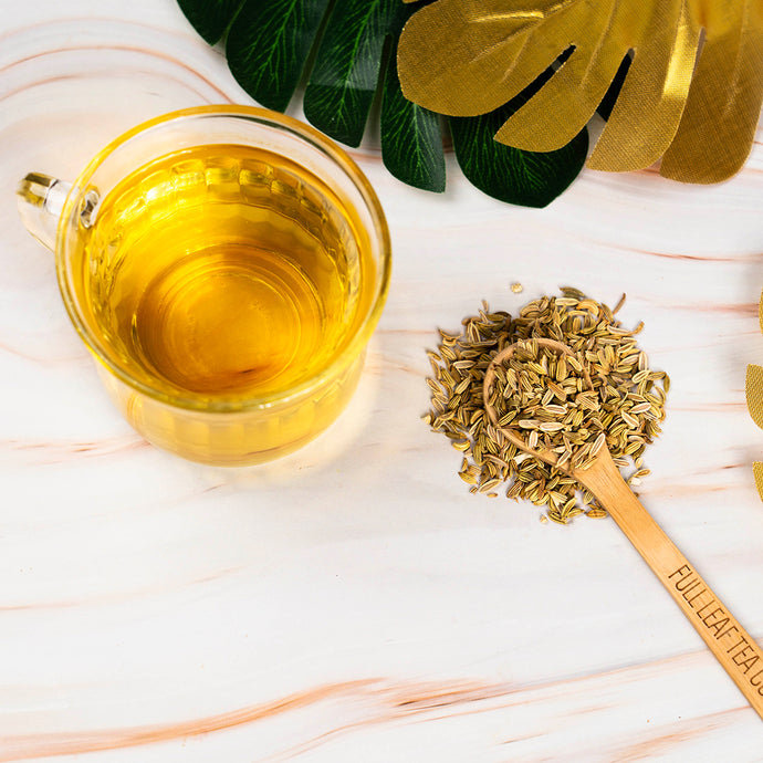 Glass mug of tea with fennel herbs on a wooden spoon, surrounded by green leaves on a light background.