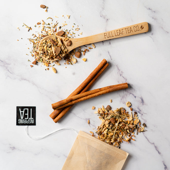 Wooden spoon with tea leaves, cinnamon sticks, and a tea bag on a marble surface with 'Full Leaf Tea Co.' branding.