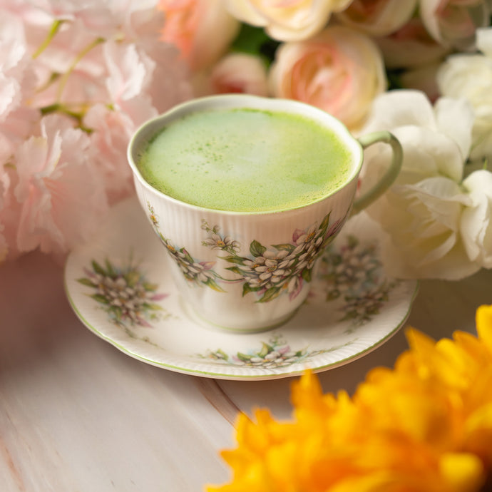 A delicate floral teacup filled with frothy Organic Culinary Matcha, placed on a matching saucer and surrounded by soft pink, white, and yellow flowers.