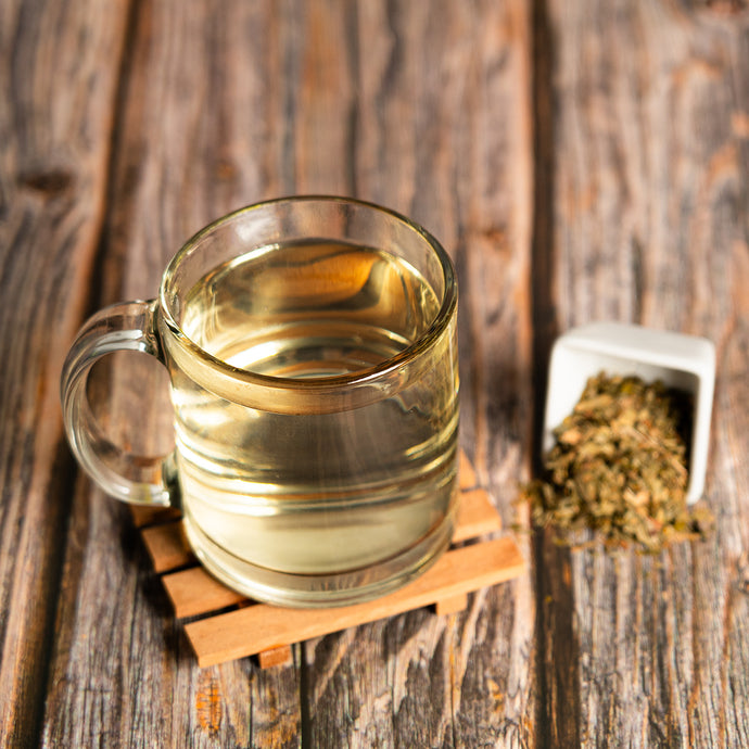 Clear mug with liquid on a wooden surface next to a small container of dried ginkgo biloba. 