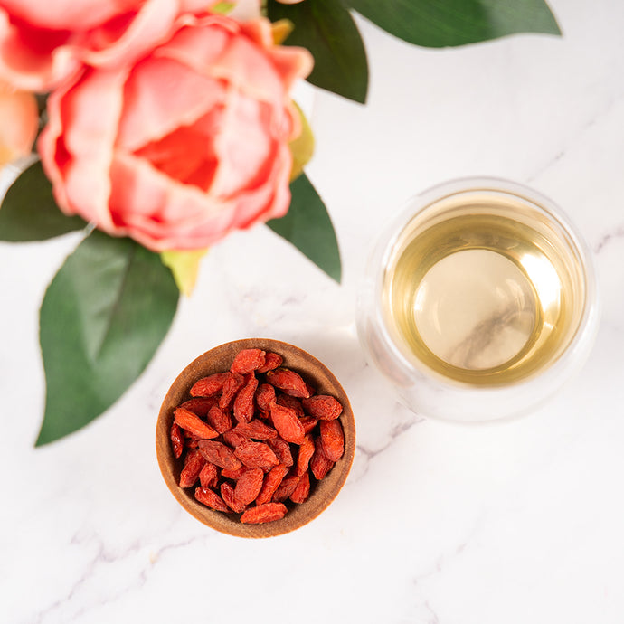 Dried goji berries in a wooden bowl with a glass of tea and flowers on a white surface