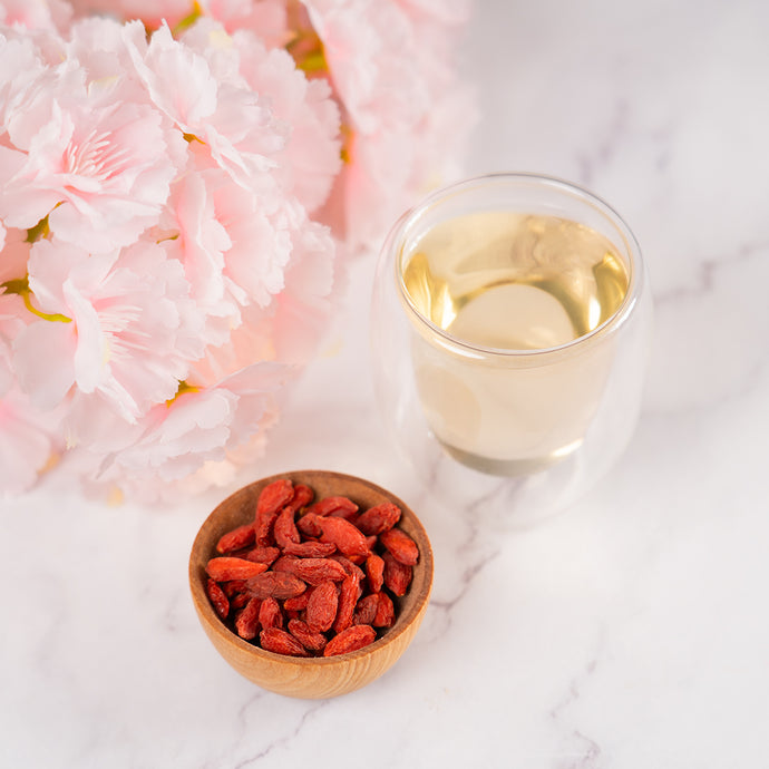 Bowl of goji berries with a glass of white tea and pink flowers on a marble surface