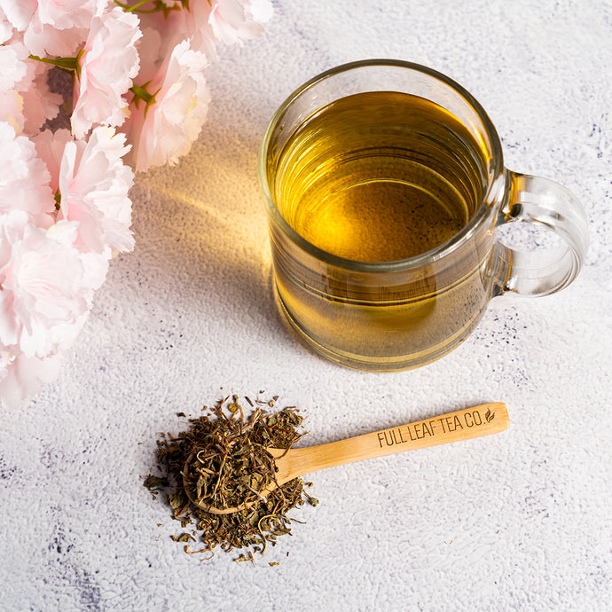 Glass mug of tea with a wooden spoon of tea leaves and pink flowers on a light surface