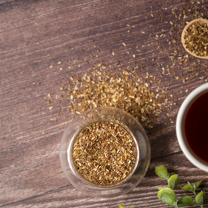 Green rooibos in a glass container on a wooden surface with a cup of tea.