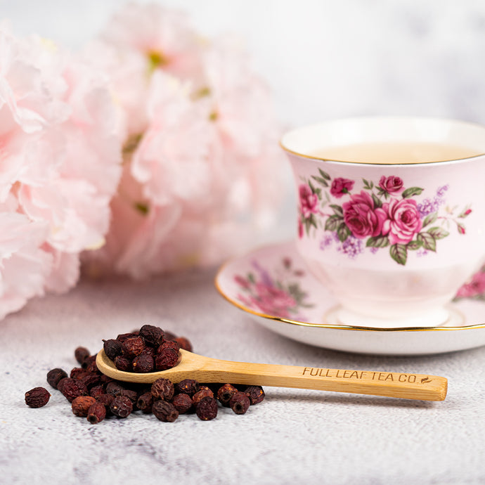 Tea cup with floral design, wooden spoon, and tea leaves on a light surface with pink flowers in the background.