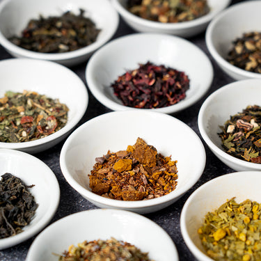 Assortment of loose-leaf teas displayed in small white bowls, featuring a variety of colorful dried herbs and tea blends on a dark surface