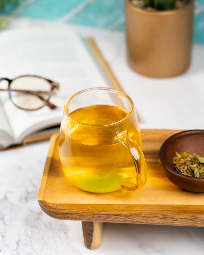Glass mug with tea on a wooden tray, next to a book and glasses on a marble surface.