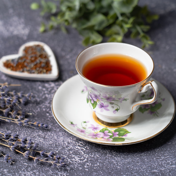 Teacup with floral design on a saucer, filled with tea, on a dark surface with lavender and heart-shaped container.