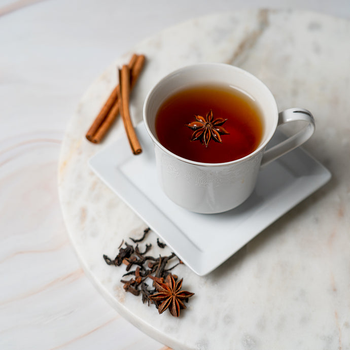 White mug of holiday cheer tea with star anise on a marble surface, surrounded by cinnamon sticks and star anise.