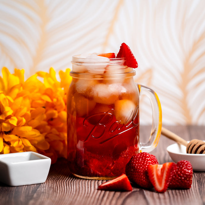 Mason jar filled with honey berry iced tea and strawberries on a wooden surface with flowers and honeycomb in the background.