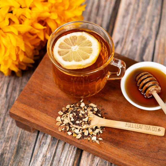 Tea cup with lemon slice, honey, and tea leaves on a wooden board with yellow flowers in the background.