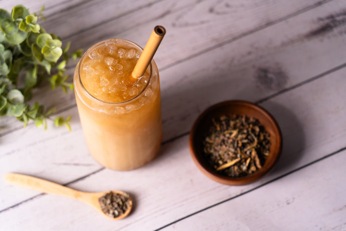 Glass of iced tea with a straw, bowl of tea leaves, and wooden spoon on a wooden surface.