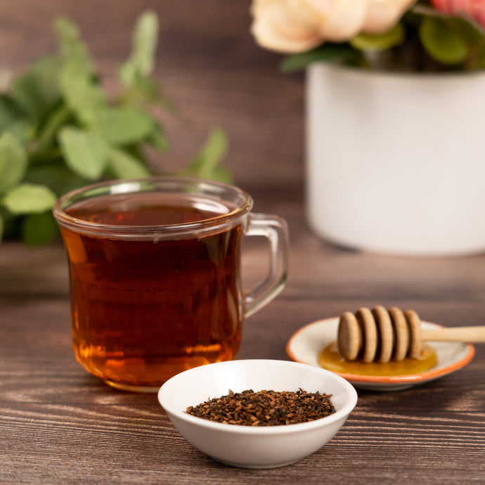 A clear mug filled with Honeybush tea with honeybush herbs in a nearby cup with flowers in the background. 