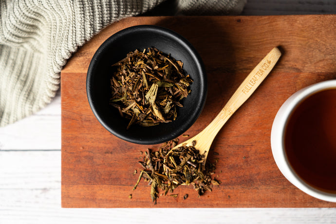 Dry houjicha tea leaves in a black bowl on a wooden tray with a wooden spoon and a cup of houjicha tea.